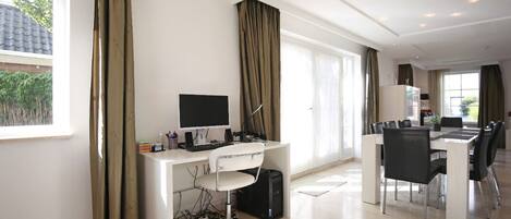 A modern office space with a desk, computer, and office chair. There is a dining area with a table and chairs.