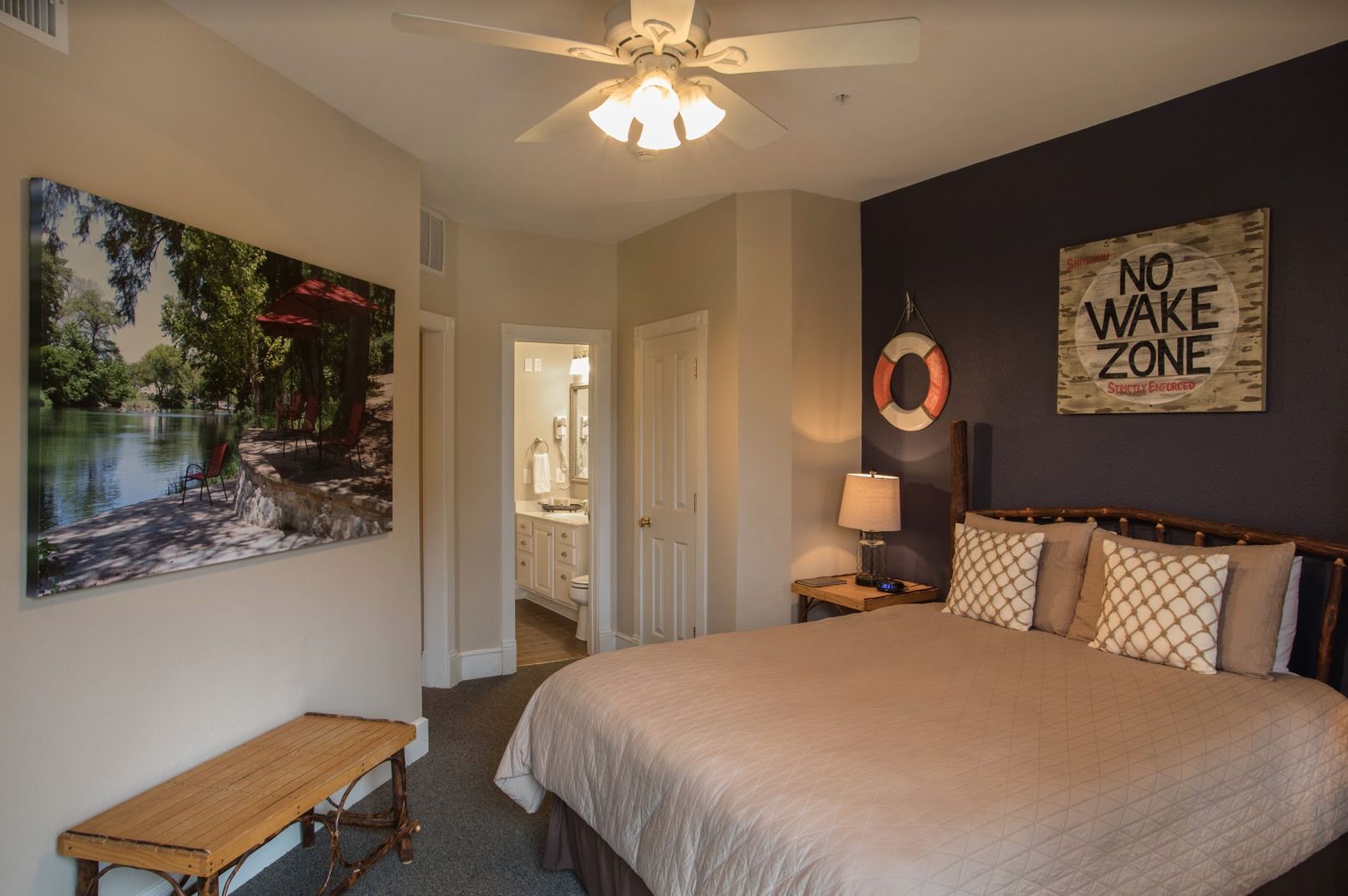 A bedroom with a large bed, a bench, a painting of a lake, a ceiling fan, and a 'No Wake Zone' sign.