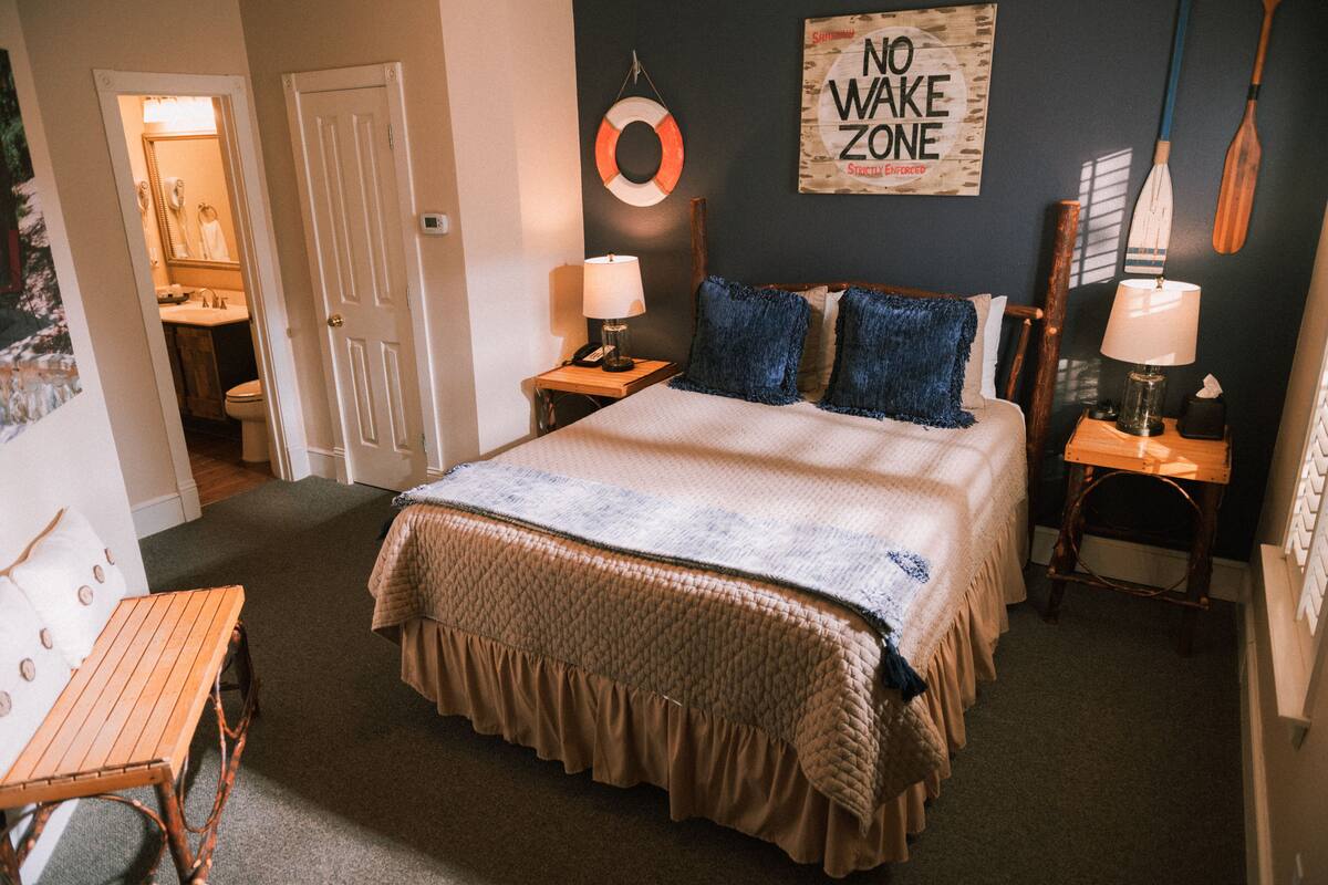 Un dormitorio con una cama grande, dos mesitas de noche con lámparas, una silla de madera, un banco, un salvavidas colgado en la pared y un letrero que dice 'NO WAKE ZONE'.