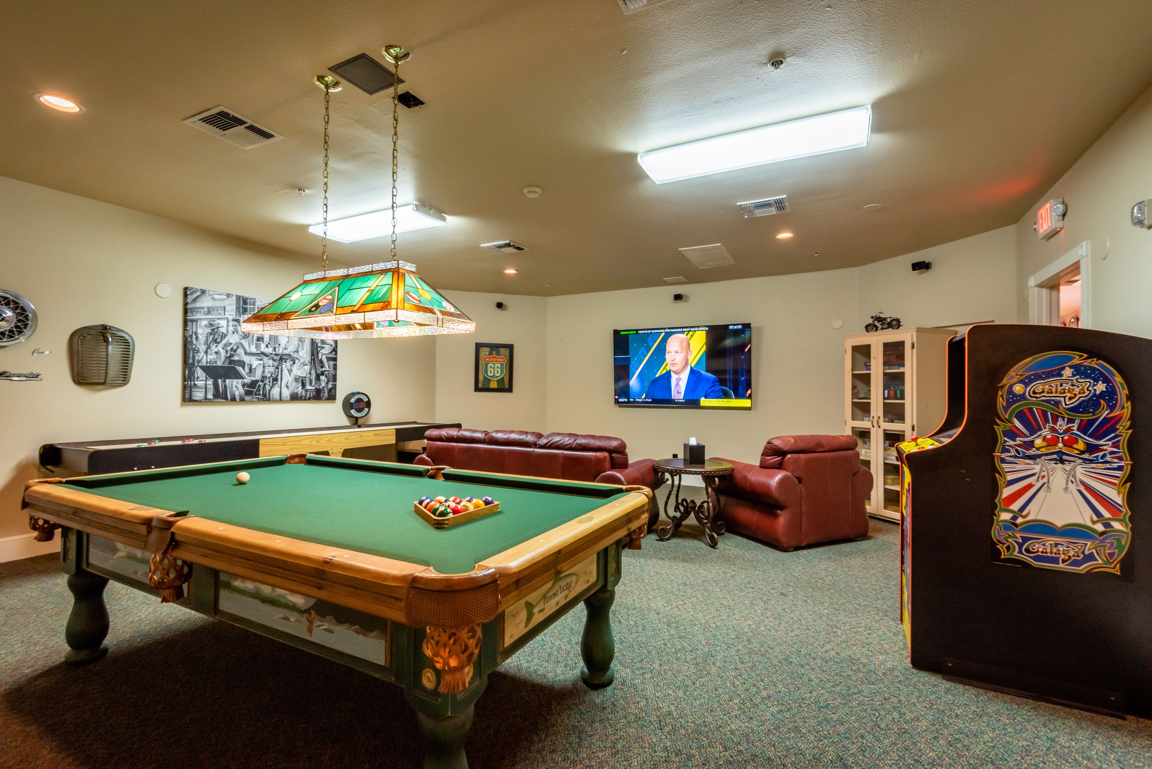 A pool table with balls, a television, a couch, and a pinball machine in a room with carpeted flooring and ceiling lights.
