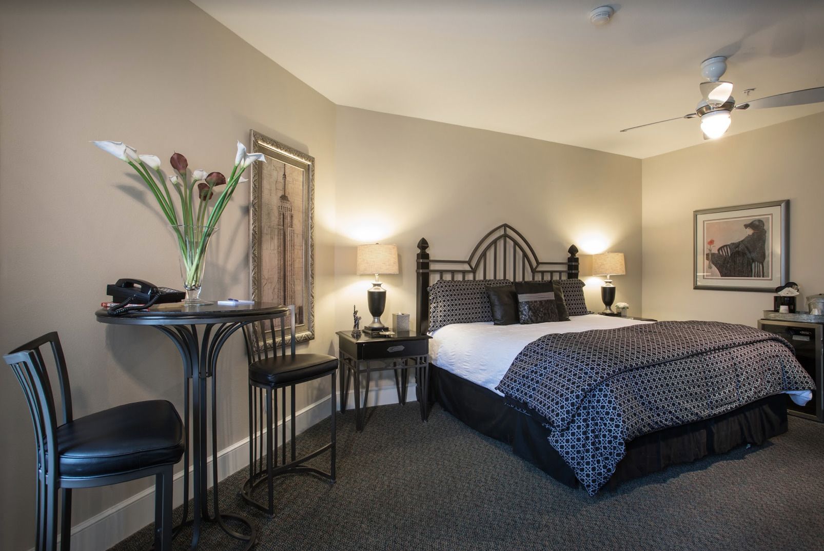 Habitación de hotel ordenada con cama, mesitas de noche, escritorio con silla y una pequeña mesa con un jarrón de flores.