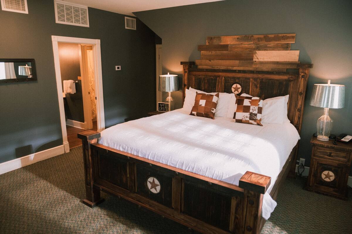 Una cama grande con cabecera de madera, dos mesitas de noche con lámparas, un espejo en la pared y una puerta que da a otra habitación.