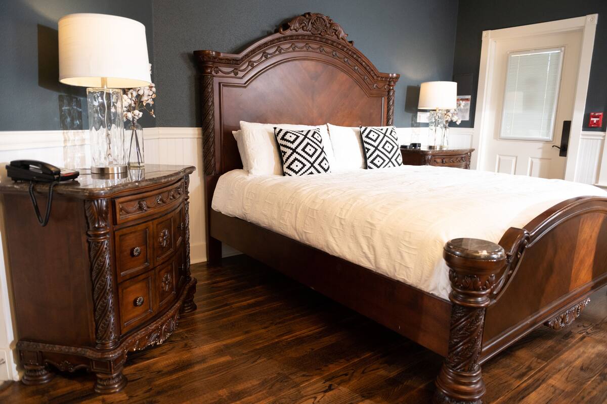 Una cama bien hecha con cabecera de madera, una mesita de noche con lámpara y un aparador con cajones.