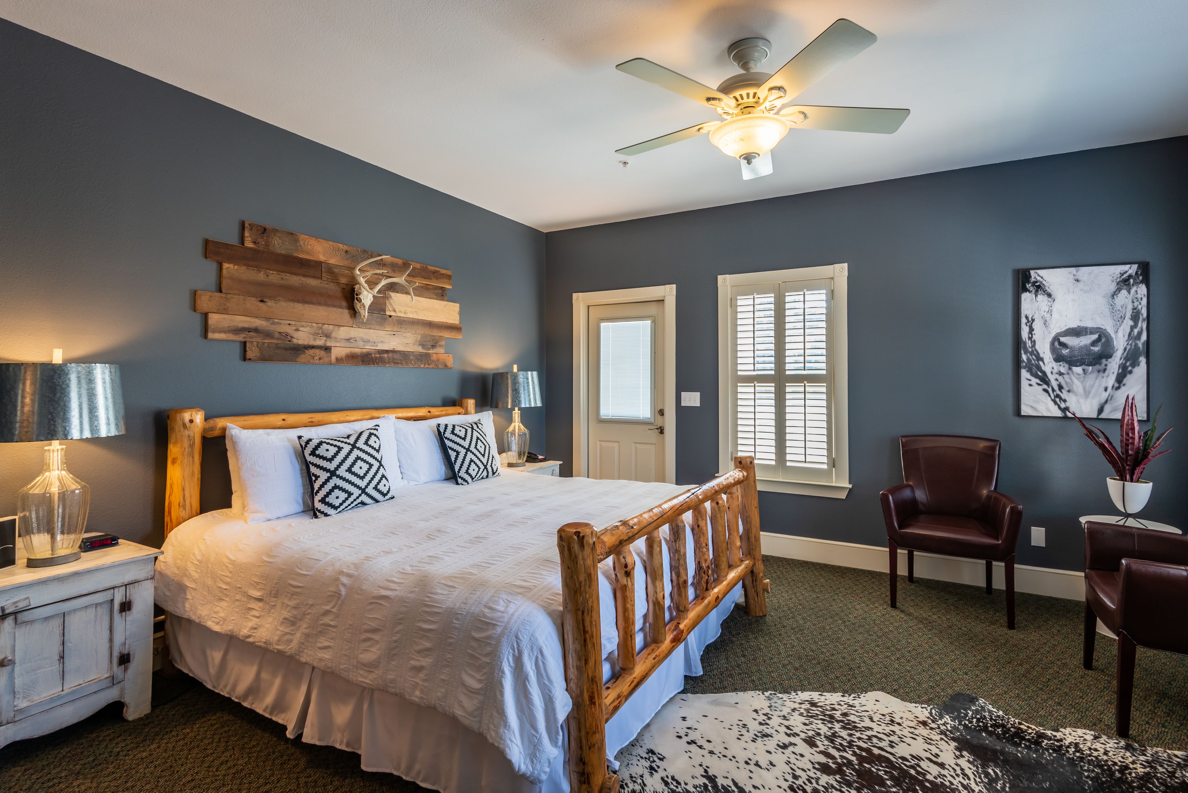 A bedroom with a wooden bed, a ceiling fan, a nightstand with a lamp, a chair, and a wall-mounted artwork.