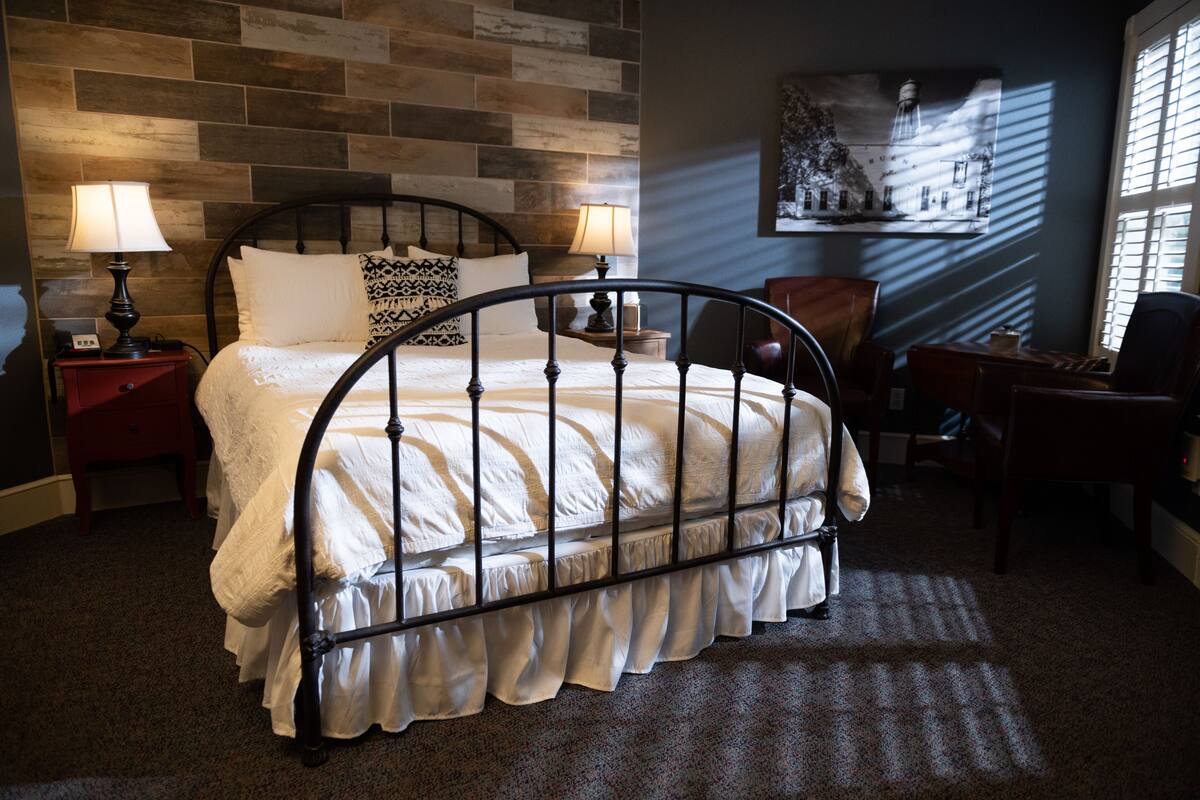 Una cama bien hecha con estructura metálica, dos mesitas de noche con lámparas, una silla y una obra de arte en la pared.