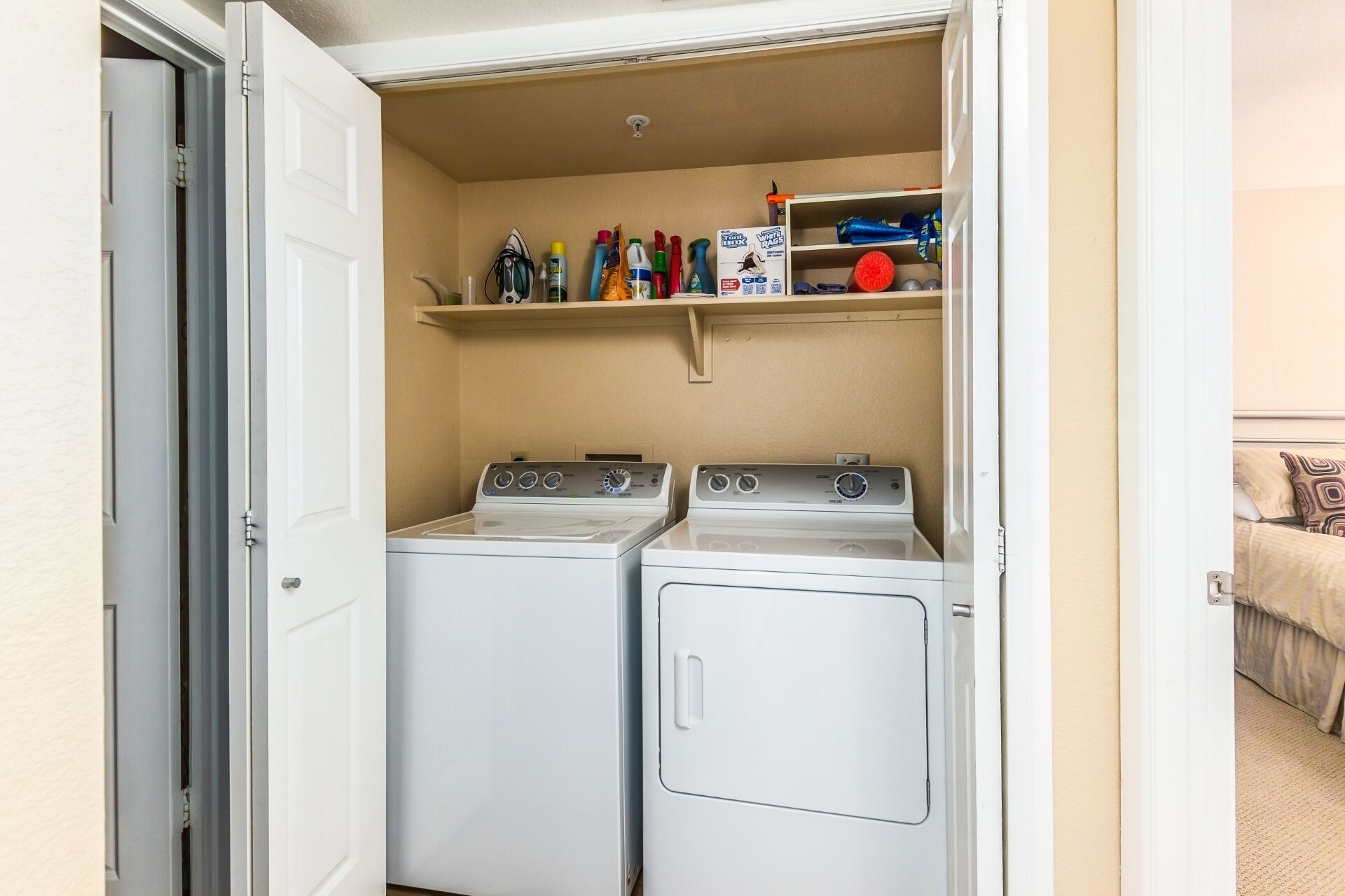 A laundry room with two front-loading washing machines and a shelf above containing cleaning supplies.