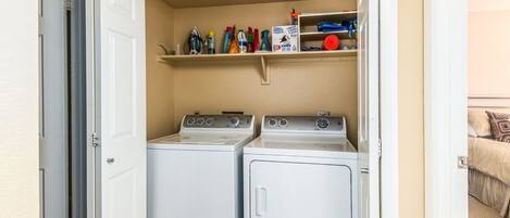 A laundry room with two front-loading washing machines and a shelf above containing cleaning supplies.