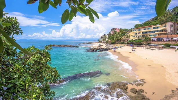 Une plage à l’eau turquoise et limpide, un rivage sablonneux et des bâtiments dotés de balcons donnant sur la mer.