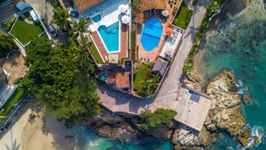 Vue aérienne d’une propriété côtière dotée de deux piscines, d’une plage et d’un terrain rocheux.