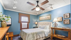 Une chambre à coucher comprenant un lit, un bureau, une chaise, un ventilateur de plafond et une fenêtre avec des stores.