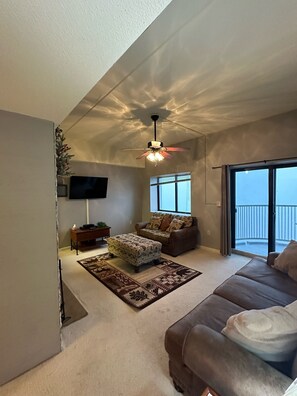 A spacious living room with a ceiling fan, a sofa, an ottoman, a TV, and a large window with a view. - 2nd Floor 'Sky Rise Vista' - Pool Access! (Sugar Mountain)
