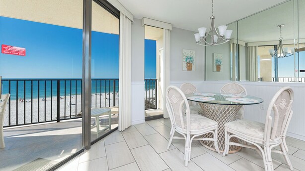 A balcony with a glass door, a round glass table with wicker chairs, and a view of the beach. - Charming One Bedroom Condo at Phoenix V Unit #411 (Orange Beach)