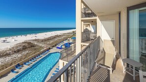 A pool with blue water, a beach with white sand, and a balcony with a table and chairs. - Charming One Bedroom Condo at Phoenix V Unit #411 (Orange Beach)
