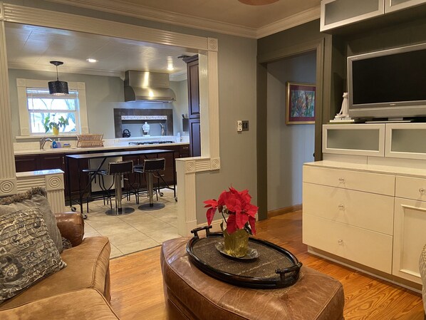 A modern kitchen with a central island, a dining area with bar stools, and a living room with a leather sofa and a decorative tray.
