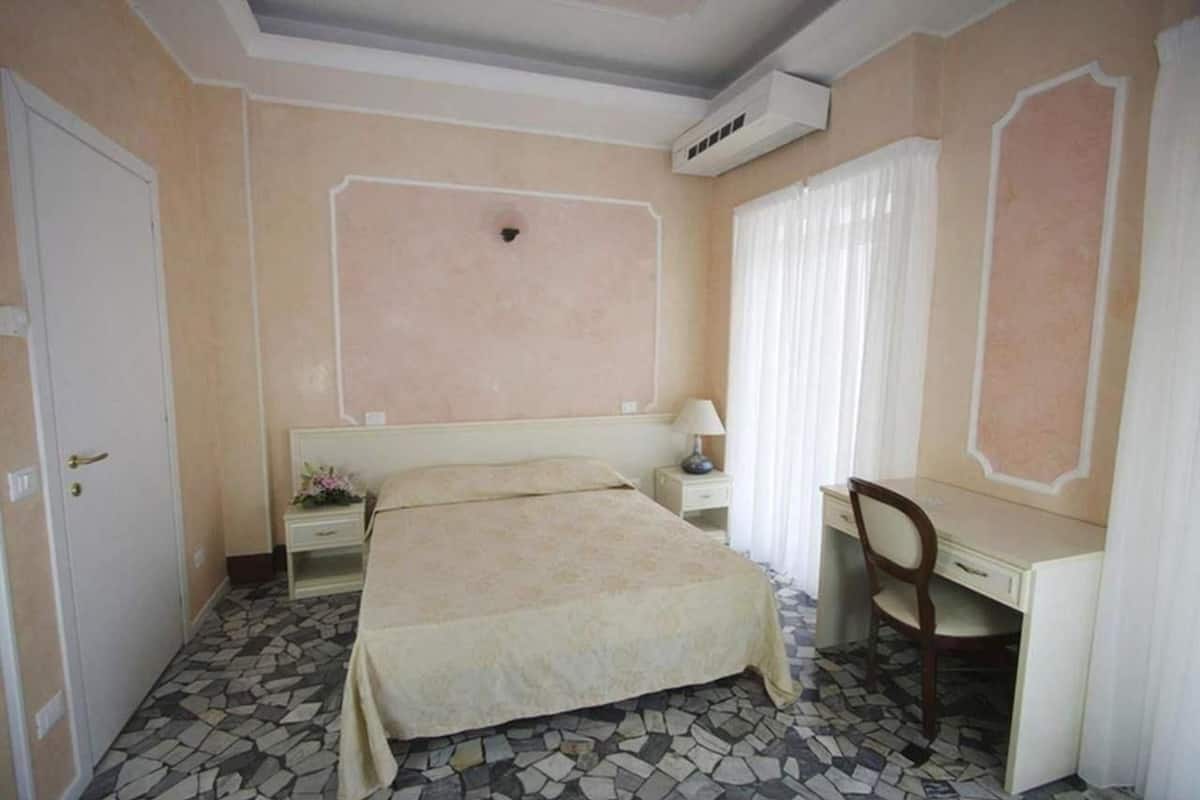 Een slaapkamer met een bed, een bureau, een stoel, een nachtkastje en een raam met gordijnen.