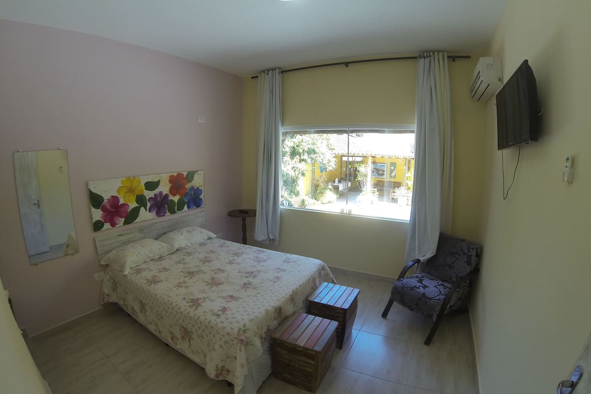 Een slaapkamer met een bed, een stoel, een klein tafeltje, een raam met gordijnen en een airconditioning.