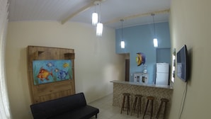Een woonkamer met een houten kast, een bank en een schilderij van vissen. Er is een keukenhoek met een koelkast, een spoelbak en een televisie aan de muur bevestigd.