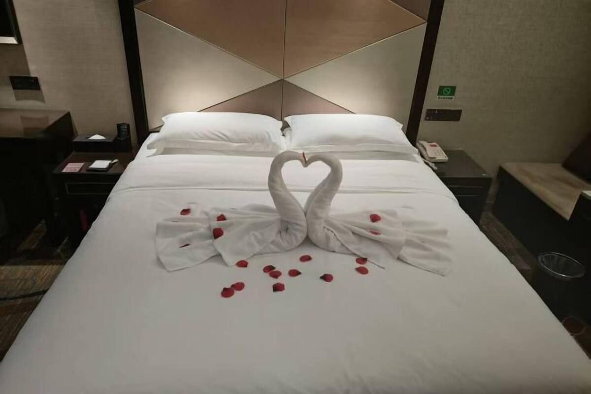 Ein Hotelzimmer mit einem ordentlich bezogenen Bett, einer Handtuch-Herzen-Arrangement und einem Nachttisch mit Lampe und Telefon.