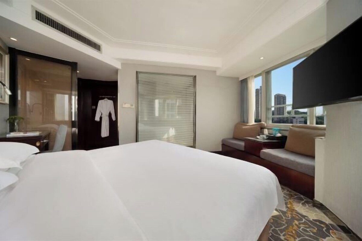 A hotel room with a large bed, a seating area with a sofa and armchairs, a television, and a window with a city view.
