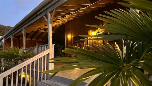 A wooden deck with a railing, leading to a covered porch with wooden beams and a light fixture. - Atea Lodge (Coromandel)