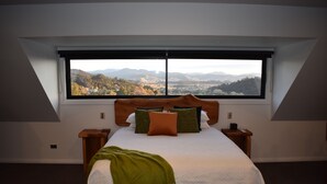 A bedroom with a large window, a bed with pillows, a bedside table, and a blanket. - Atea Lodge (Coromandel)