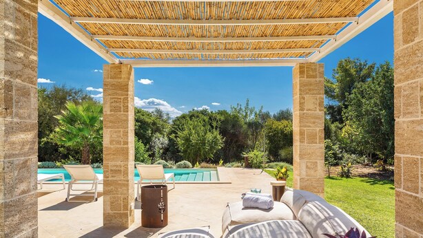 A covered outdoor seating area with stone pillars, a pool in the background, and a lush garden. - Villa Chiara by Perle di Puglia (Ruffano)