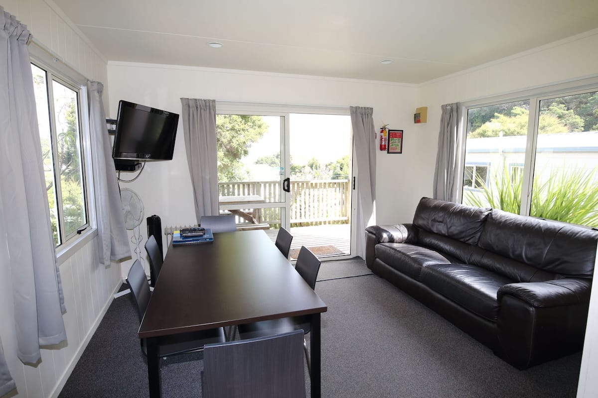 A room with a dining table, chairs, a black leather sofa, a flat-screen TV mounted on the wall, and a sliding glass door leading to an outdoor area.