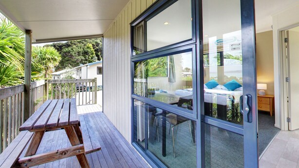 A wooden deck with a table and chairs, a glass door leading to a room with a bed and a nightstand, and a view of the outdoors. - Russell Top 10 Holiday Villas (Russell)
