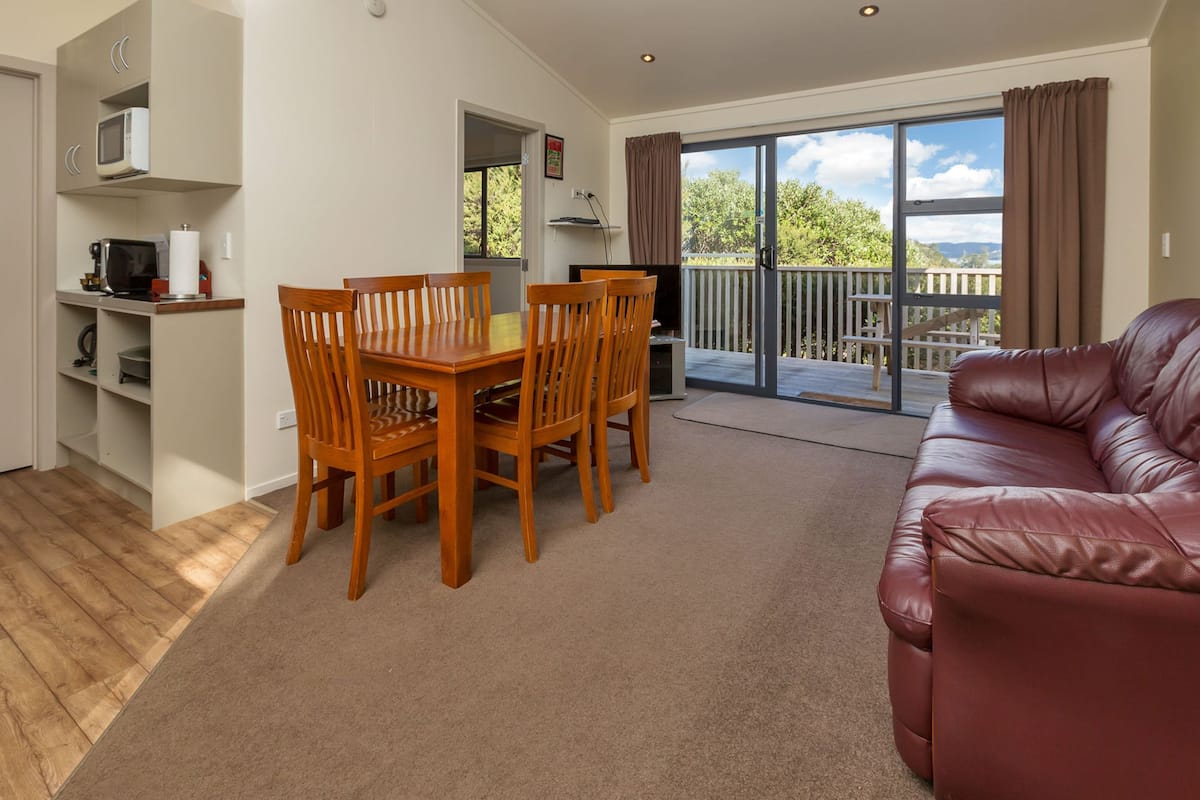 A living room with a wooden dining table, chairs, a red leather sofa, a TV, and a balcony with a view of trees and hills.