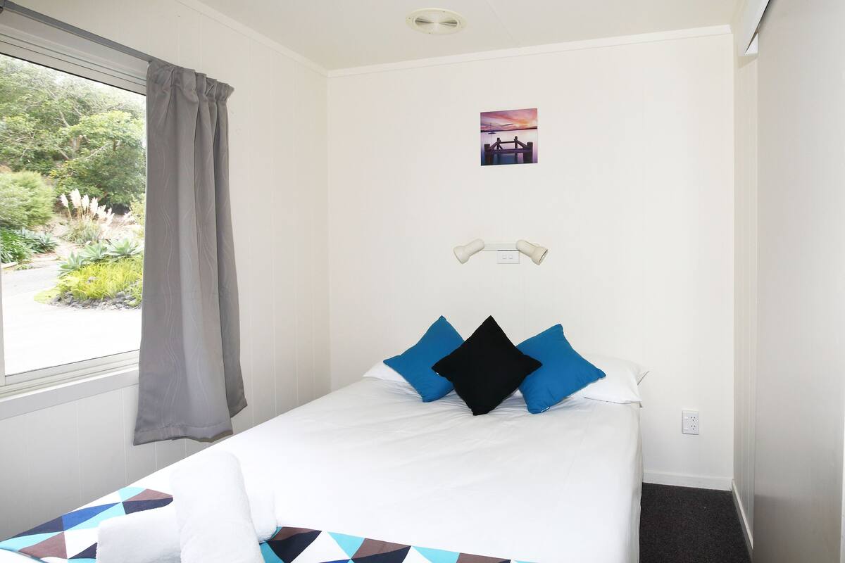 A neatly made bed with a geometric-patterned bedspread, a window with a view of greenery, and a small picture on the wall.