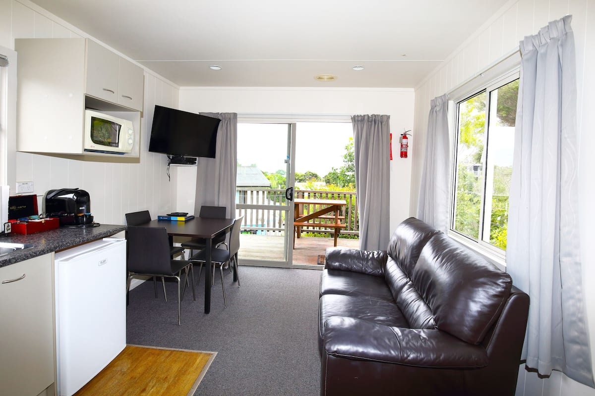 A compact kitchen with a microwave, a coffee maker, and a small refrigerator. There is a dining area with a table and chairs, and a leather sofa near a large window with curtains.