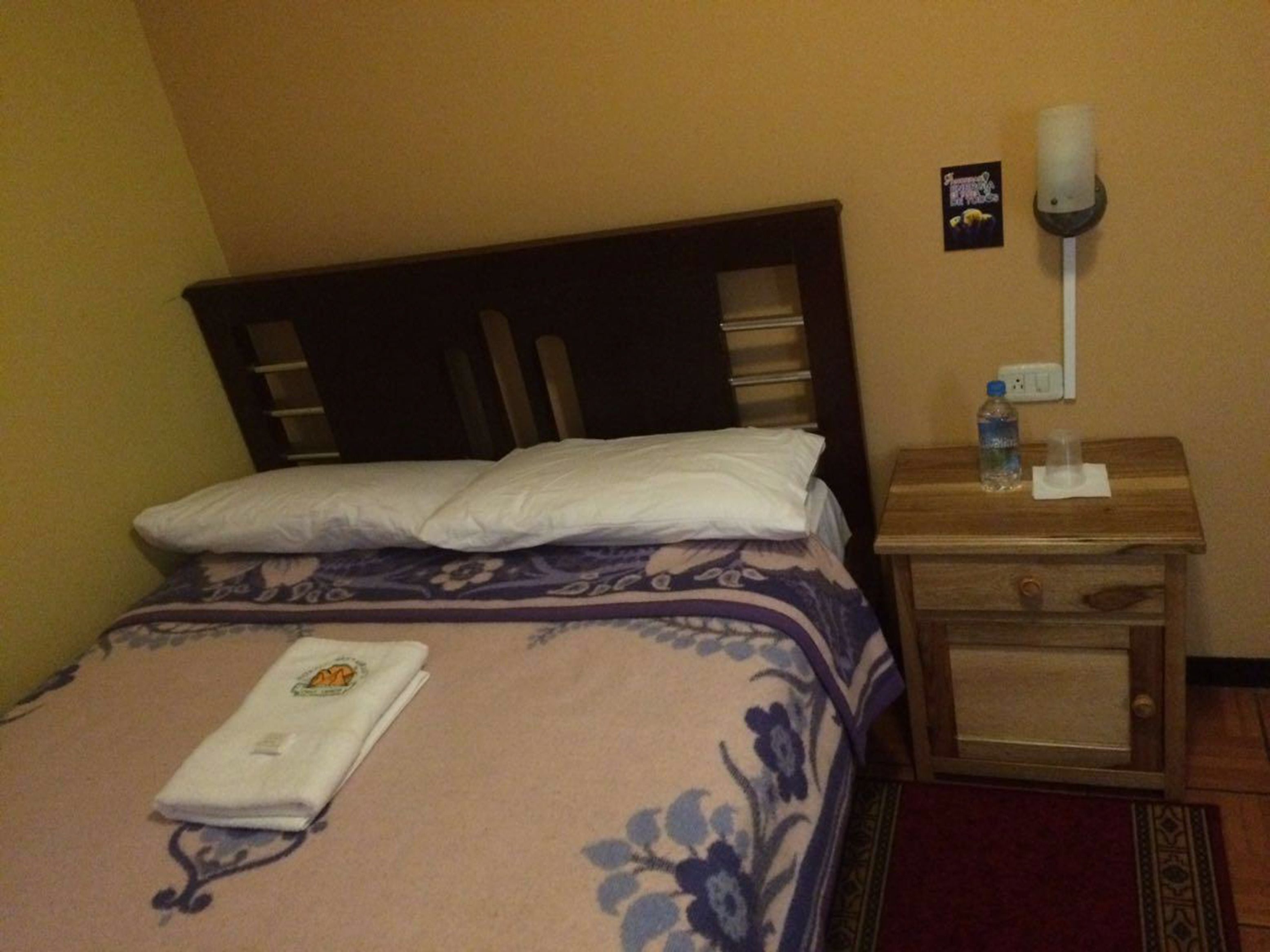 Una habitación de hotel con una cama, una mesita de noche de madera, una botella de agua y una toalla.