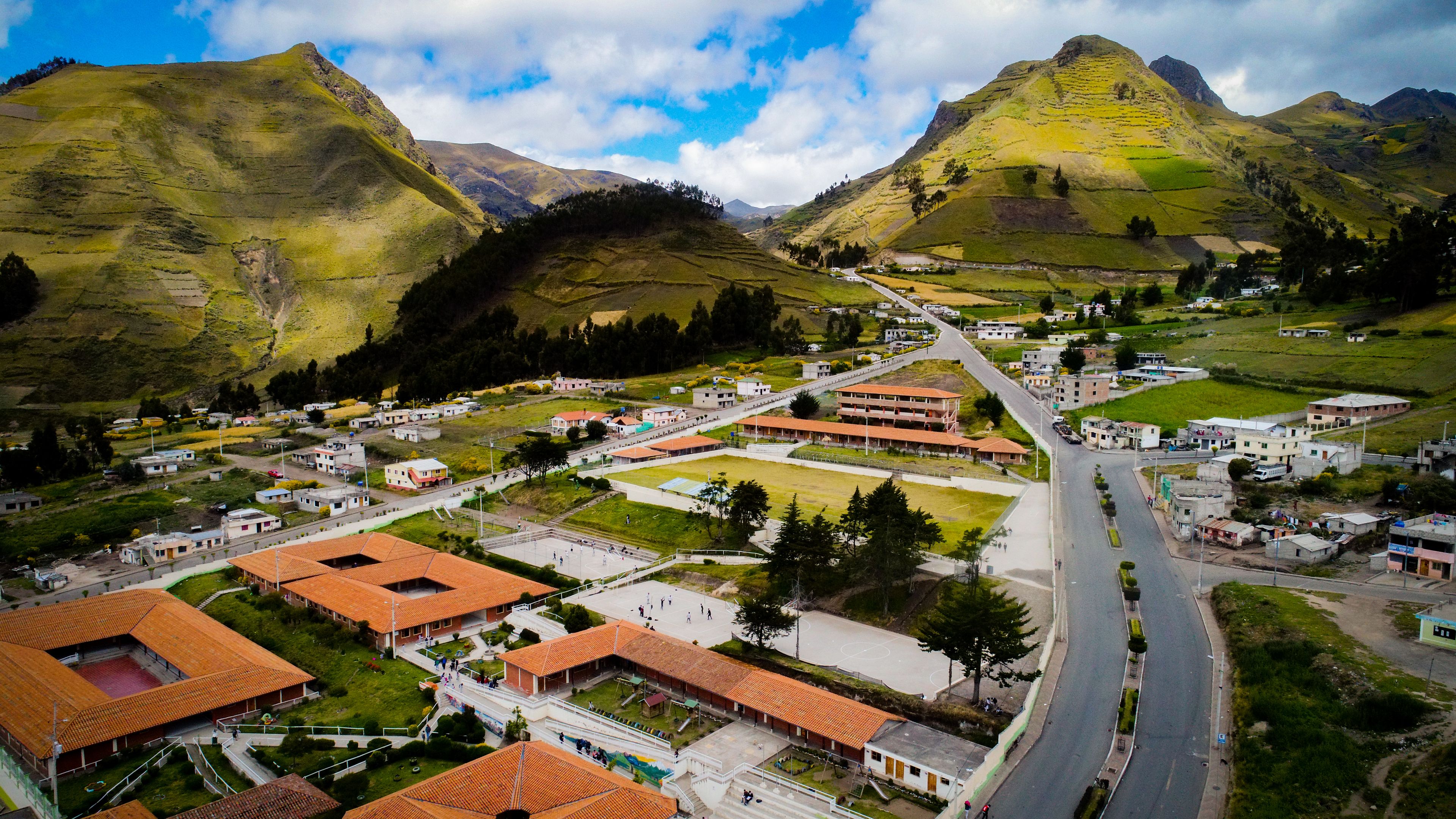 Un pueblo con una calle principal, edificios con tejados de tejas rojas y colinas verdes al fondo.