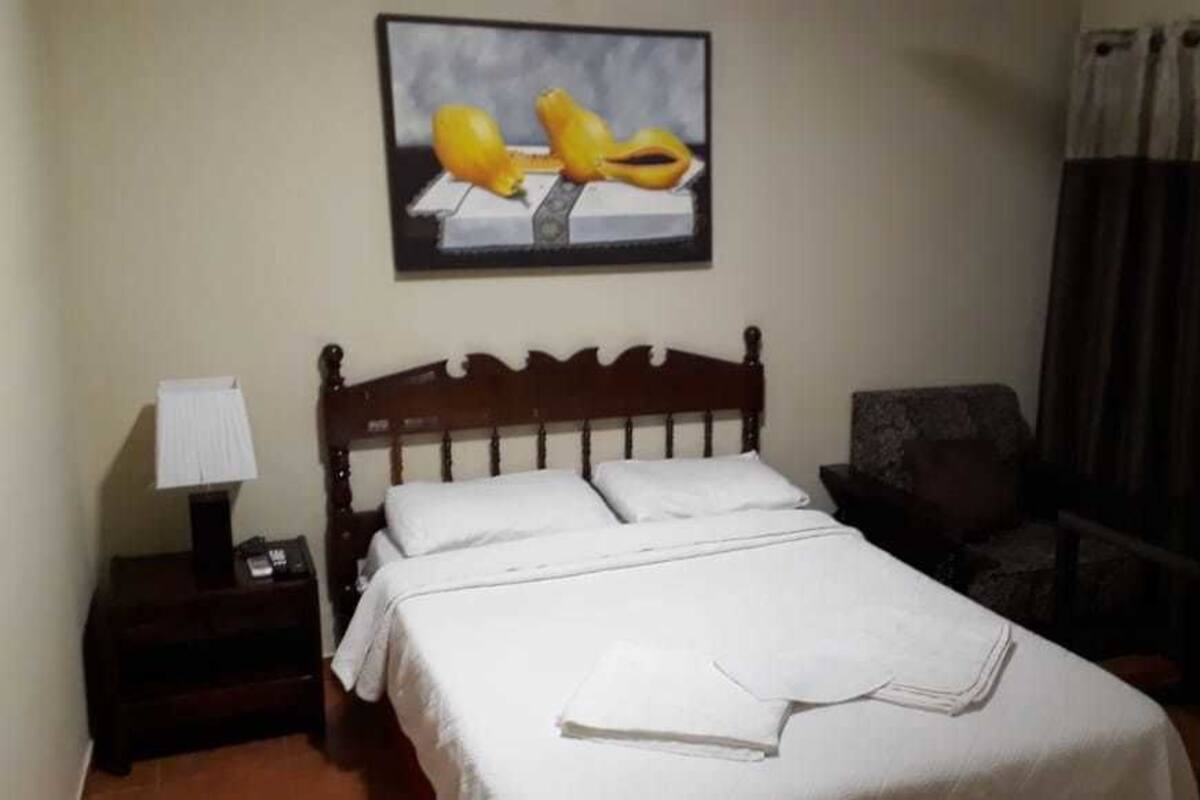 A hotel room with a wooden bed, a nightstand with a lamp, a chair, and a painting on the wall.