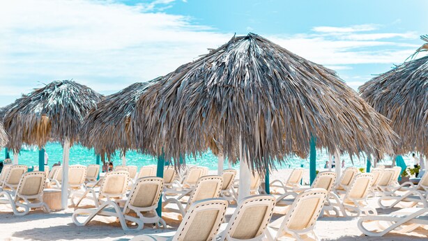 Sillas de playa dispuestas bajo sombrillas de paja en una playa arenosa.