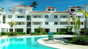 Un resort moderno con una piscina central, varias edificaciones blancas con techos de tejas rojas y abundante follaje.