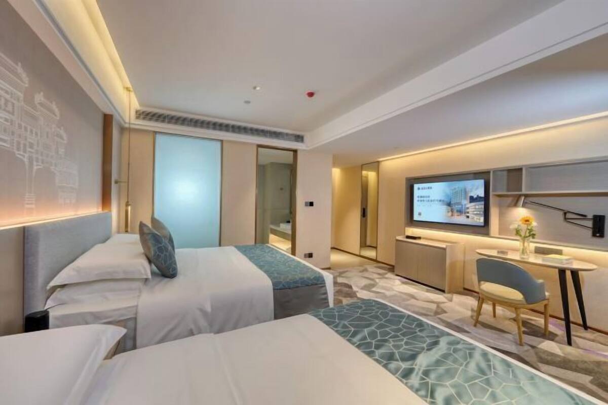 A hotel room with two beds, a desk, a chair, a TV, and a small table with a vase.