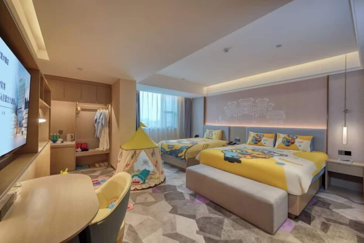 A hotel room with two beds, a TV, a desk, a chair, a small play tent, and a large window.