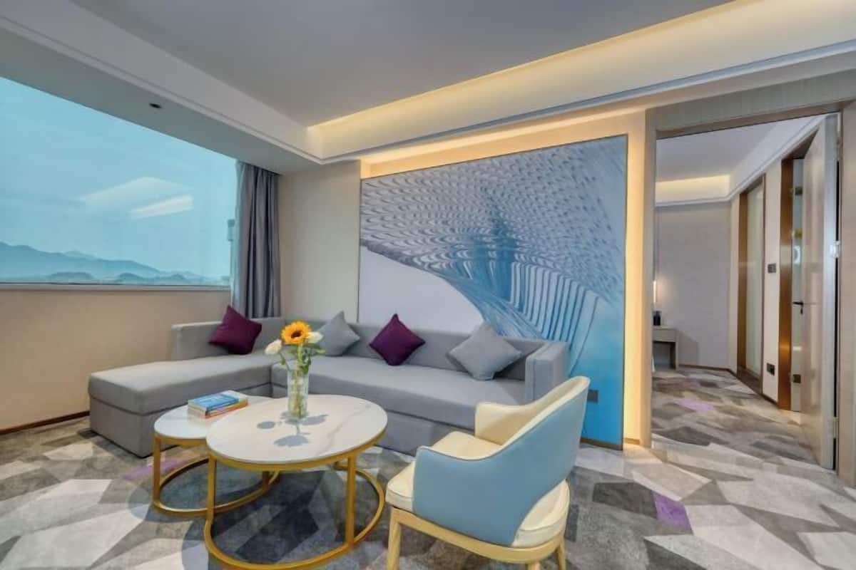 A modern hotel room with a sofa, two armchairs, a round table, and a decorative wall piece.