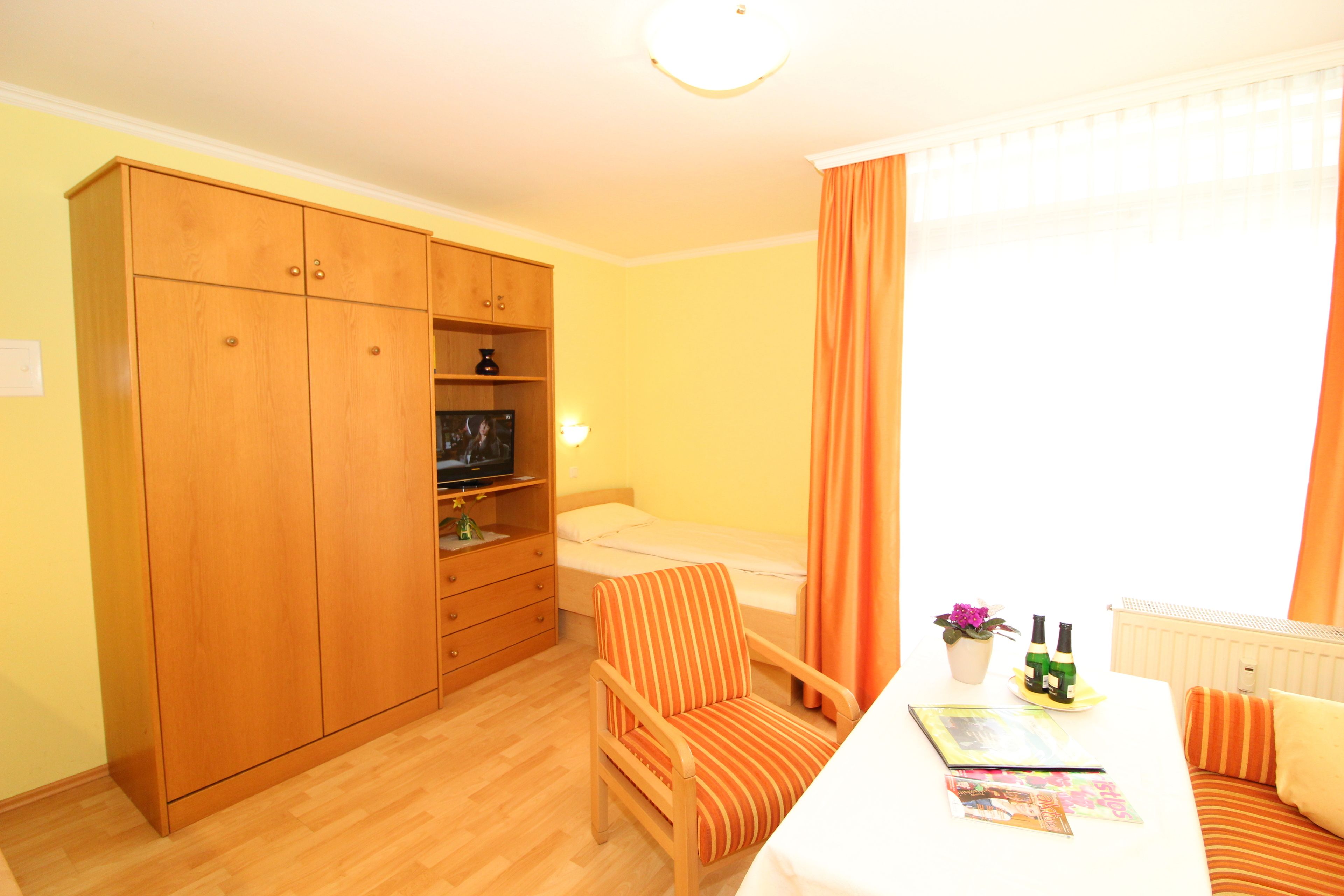 Une petite pièce bien éclairée, avec un lit simple, une armoire en bois, un fauteuil à rayures, une petite table avec un magazine et une fenêtre avec des rideaux.