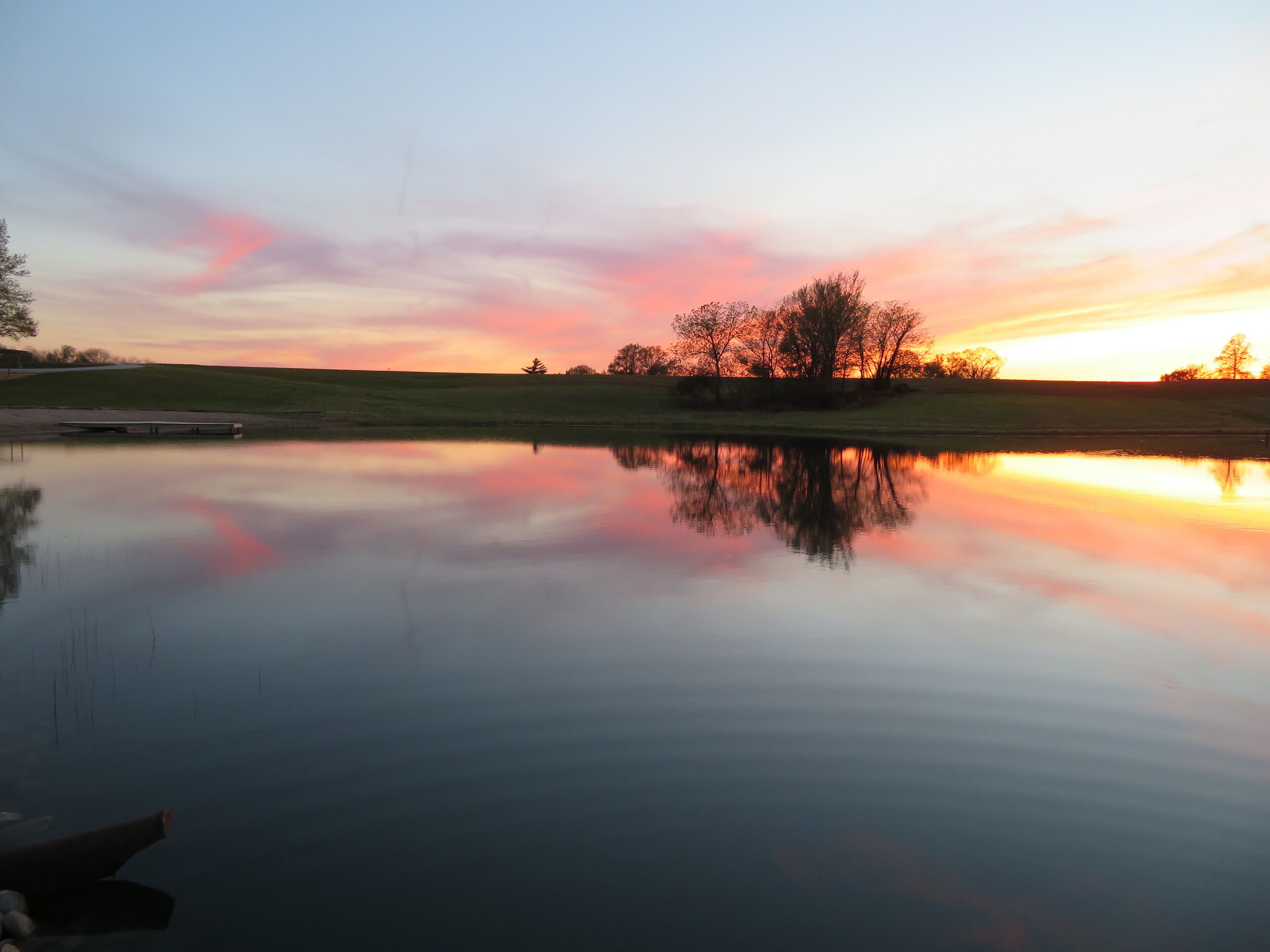 Ein ruhiger See, der einen Sonnenuntergang und Bäume am Horizont spiegelt.