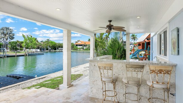 A covered outdoor bar area with three chairs, a ceiling fan, and a view of a body of water and distant buildings. -  Beautiful Waterfront Pool Home, Walk to Beach, Pool/PPong Tbl (Marco Island)