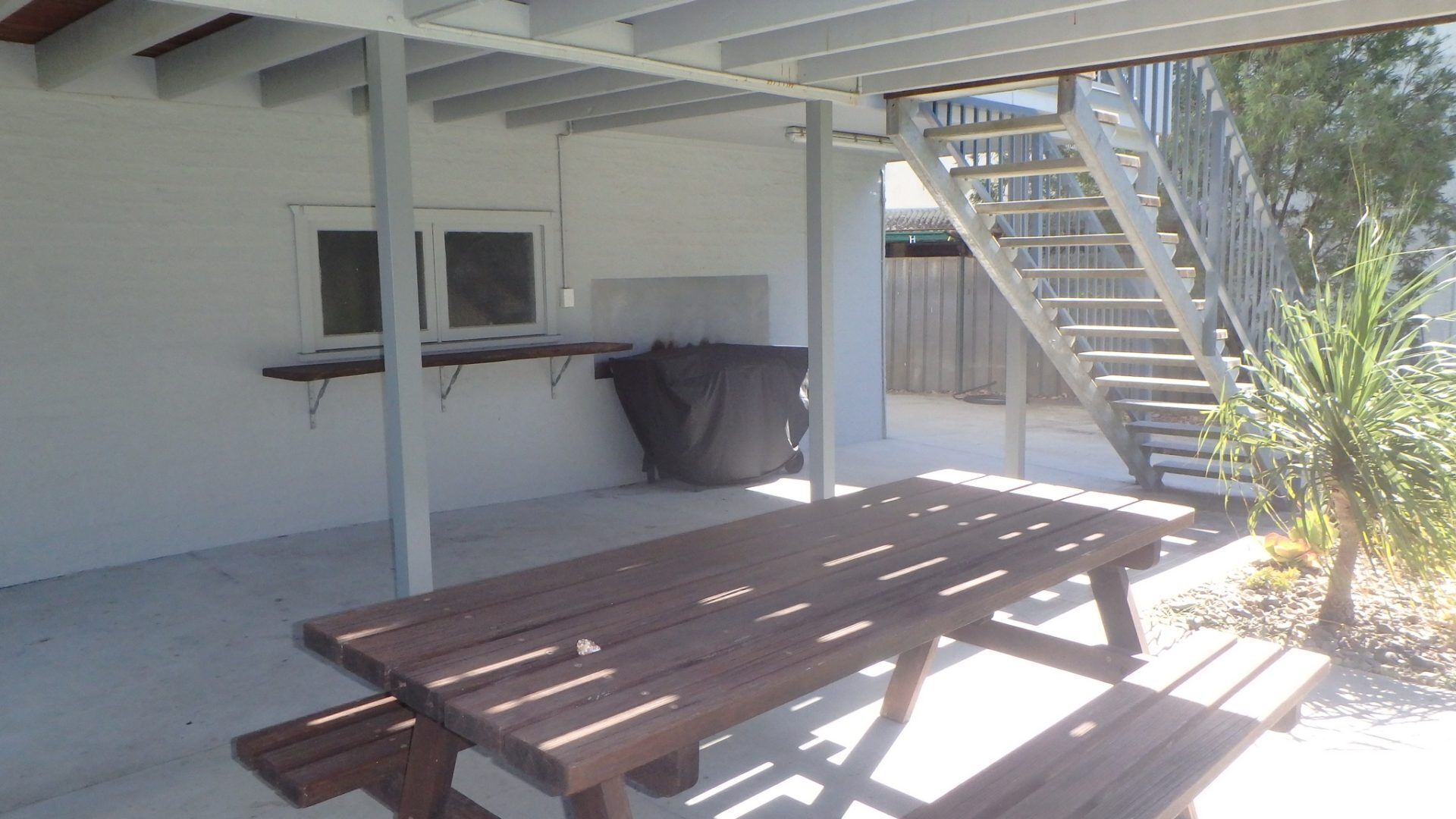Un patio couvert avec une table et des bancs en bois, une fenêtre et un escalier menant à un niveau supérieur.
