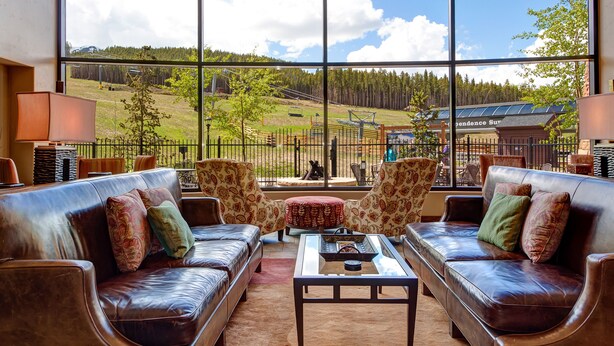 A spacious living area with leather sofas, patterned cushions, and a large window offering a view of a ski lift and forested hills. - ON THE SLOPES. FAST RESPONSE. Ski Side Corner Unit, Crystal Peak Lodge 2BR/2BA (Breckenridge)