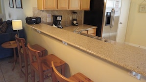 A kitchen with wooden cabinets, a granite countertop, a refrigerator, a coffee maker, and a blender. There are three wooden stools and a small table with a lamp. - Four Corners Area Pool Homes by ORB (Davenport)