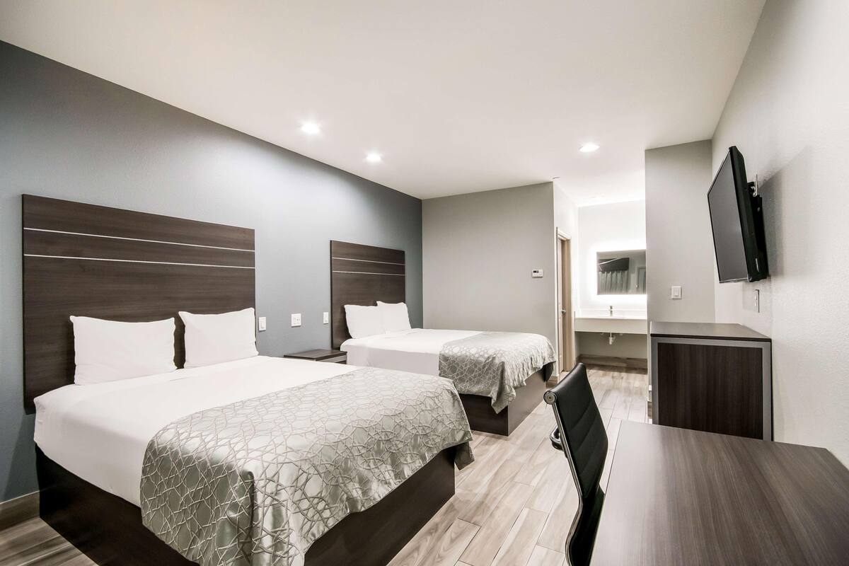 A hotel room with two beds, a desk, a chair, a TV, and a small kitchenette.