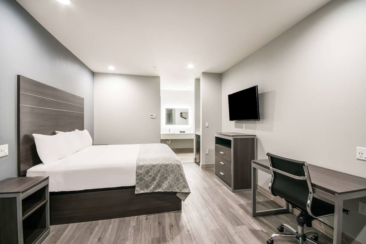 A modern hotel room with a bed, desk, chair, TV, and a bathroom in the background.