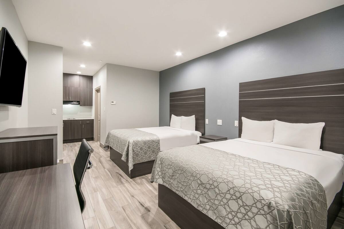 A modern hotel room with two beds, a dining area, and a kitchenette.