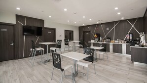 A modern, well-lit dining area with white tables and black chairs, featuring a kitchen counter with various appliances and a flat-screen TV mounted on the wall. - Americas Best Value Inn & Suites NE Houston Downtown I-610 (Houston)
