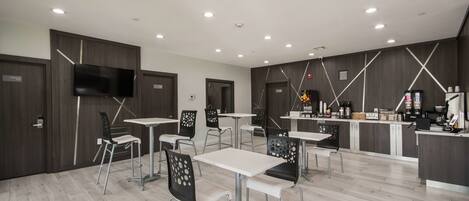 A modern, well-lit dining area with white tables and black chairs, featuring a kitchen counter with various appliances and a flat-screen TV mounted on the wall.